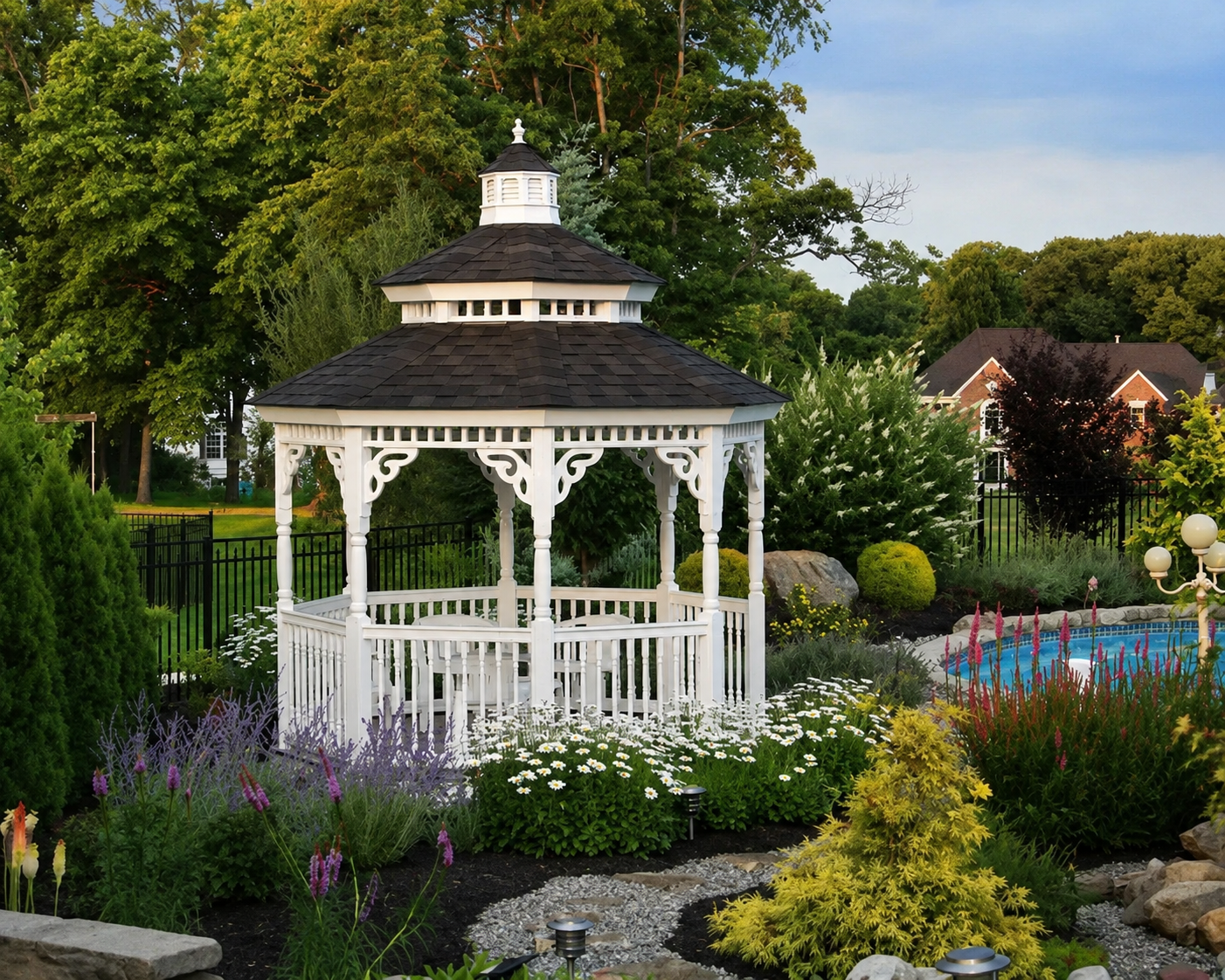 White garden gazebo surrounded by flowers, landscaping, and a backyard pool.